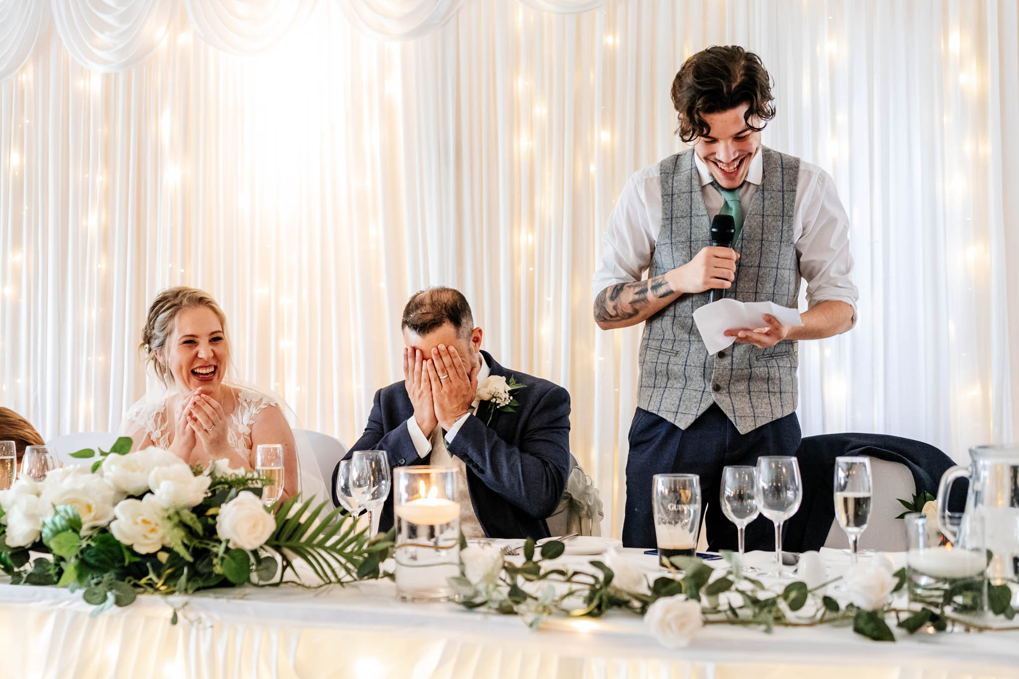 Groom covers his face with his hands as his son, the best man, tells an embarrassing story about him. The best man is holding a microphone and has a big grin on his face. The Bride is next to the groom and is laughing hard. 